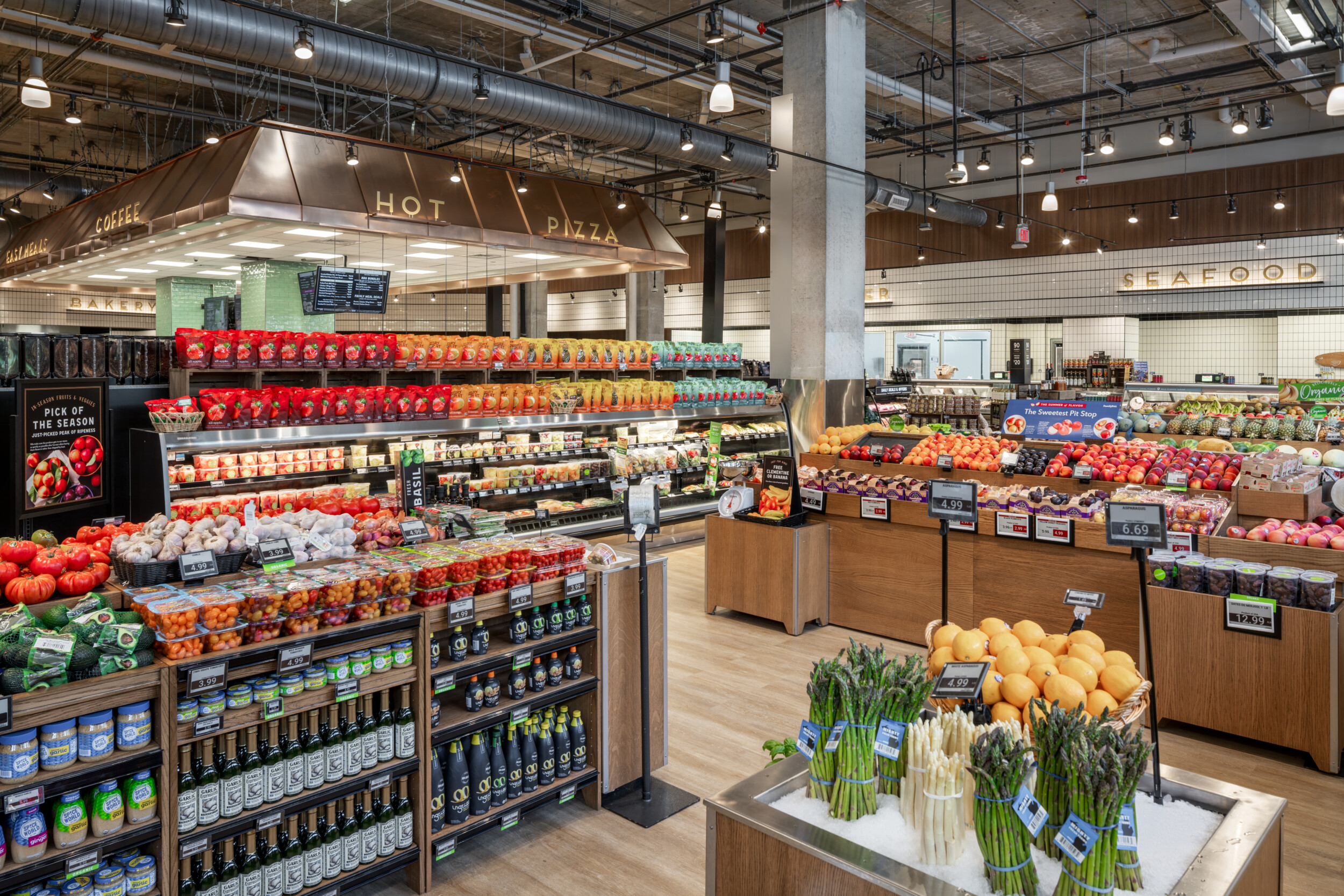 Bright, modern supermarket interior featuring fresh produce displays, refrigerated grocery items, bakery, coffee, hot pizza, and seafood departments.